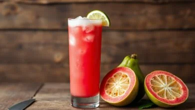 Suco De Goiaba Natural Cremoso E Refrescante Em 5 Minutos 48 Um copo alto de suco de goiaba vibrante, com espuma natural e pedaços de gelo, servido em fundo de madeira rústica.